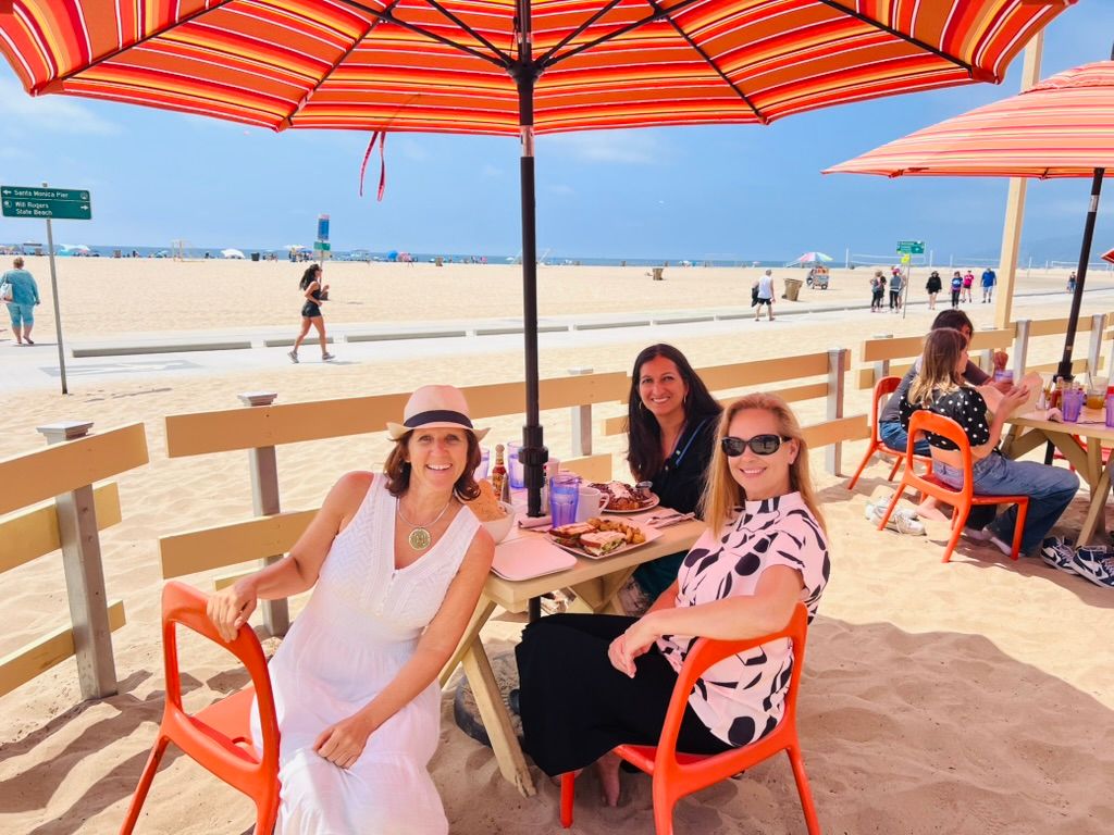 Claudia in a white dress and hat with two friends at a table on the sand under striped umbrellas, Santa Monica beach behind them