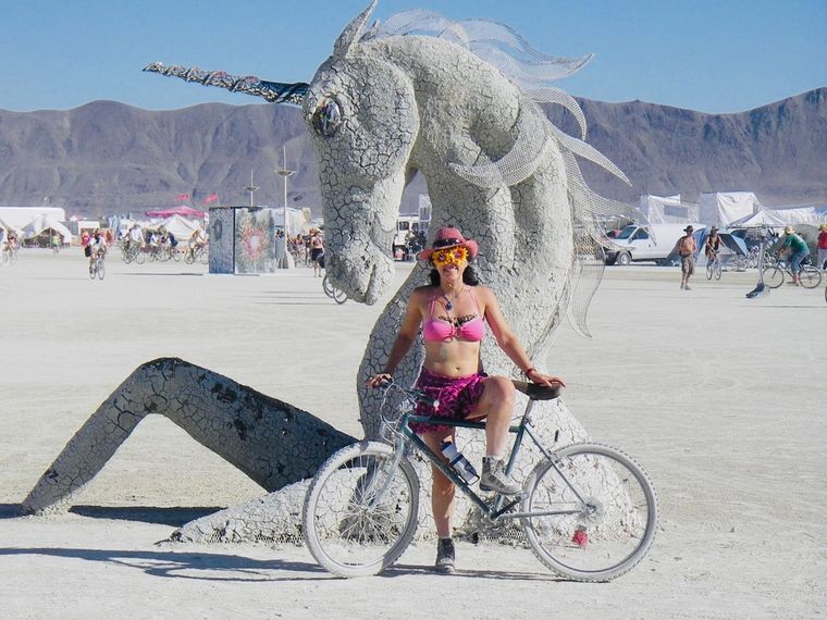 Claudia on a bicycle in a cowboy hat and sunflower sunglasses beside a giant unicorn sculpture on the playa