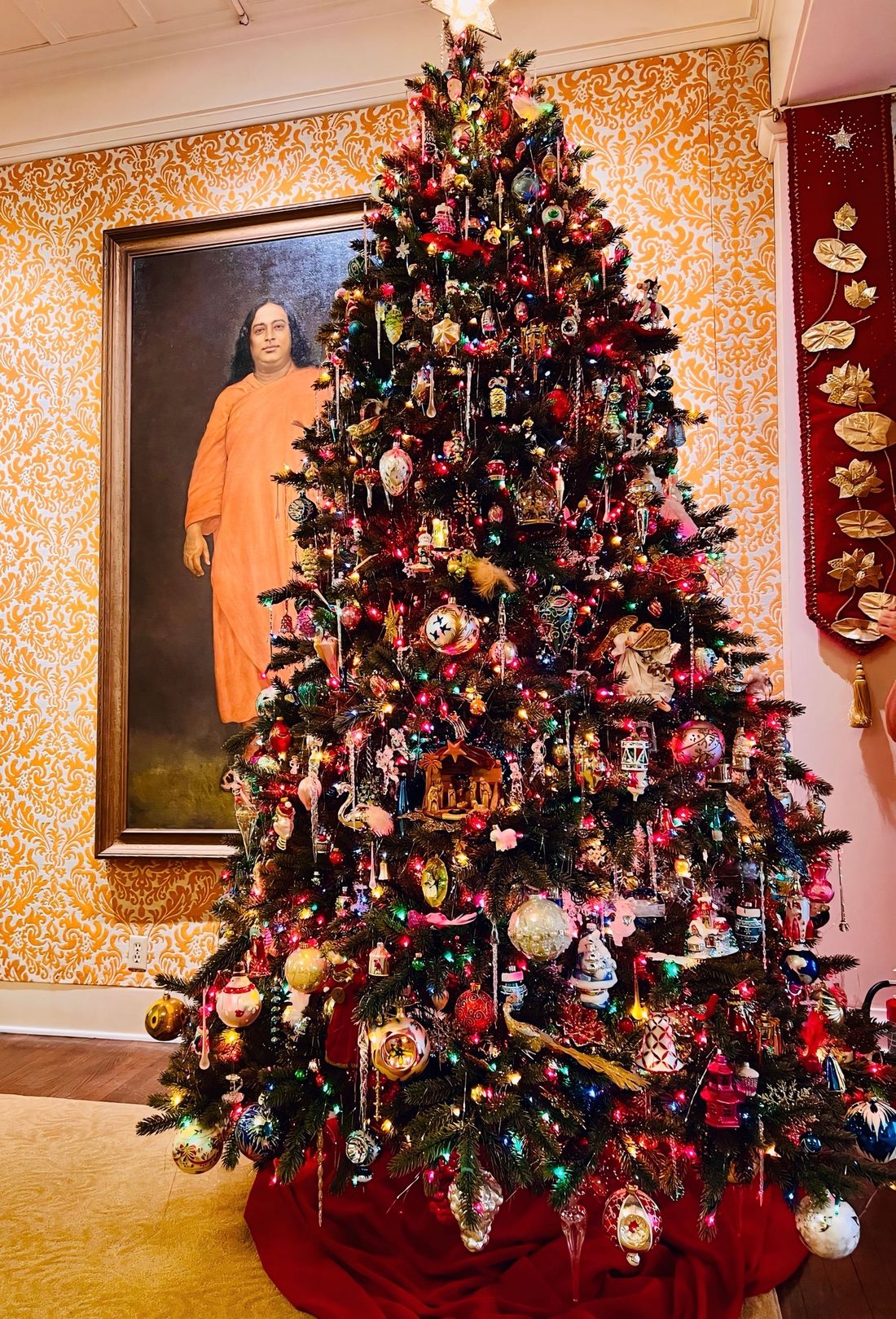 A Christmas tree crowded with colorful vintage ornaments in front of a large portrait of a spiritual teacher in an orange robe, on gold damask wallpaper