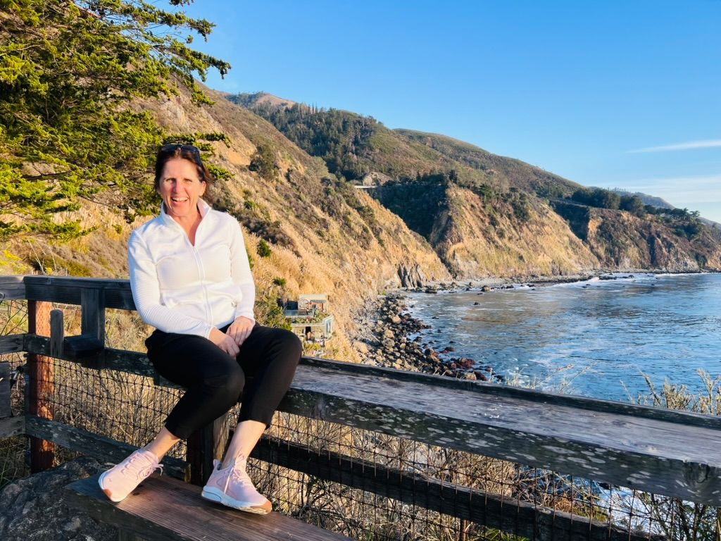 Claudia smiling on a wooden fence above a rocky stretch of the California coast, golden hills behind her