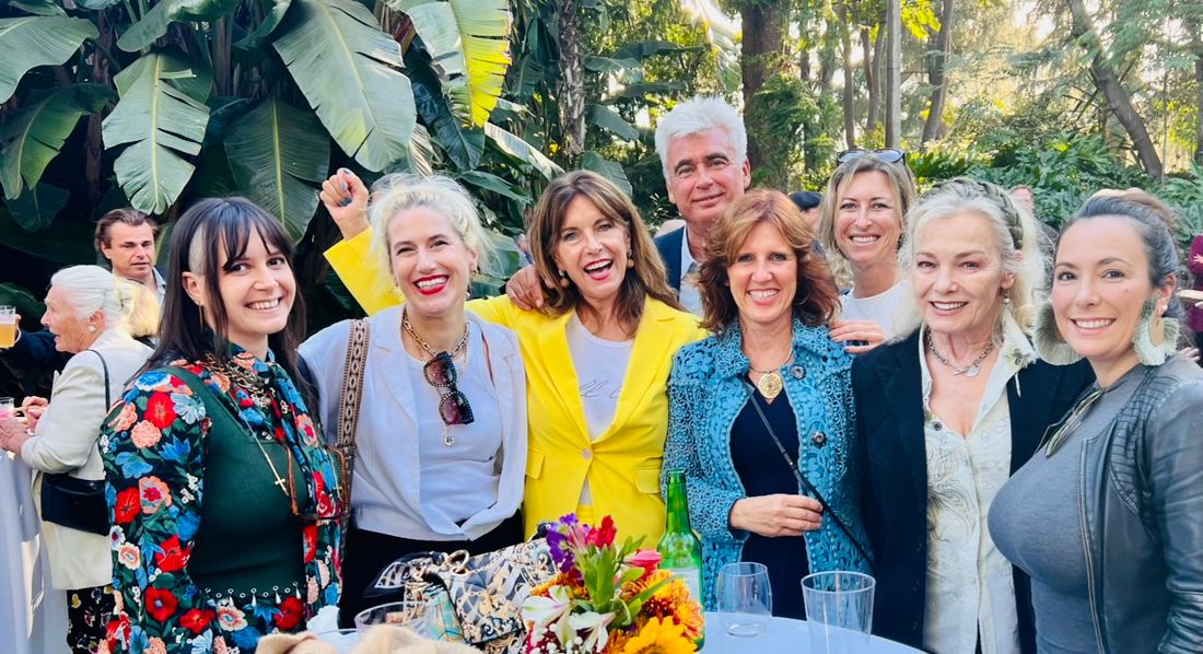 Claudia laughing with a group at an outdoor garden reception, banana palms behind them and flowers on the table