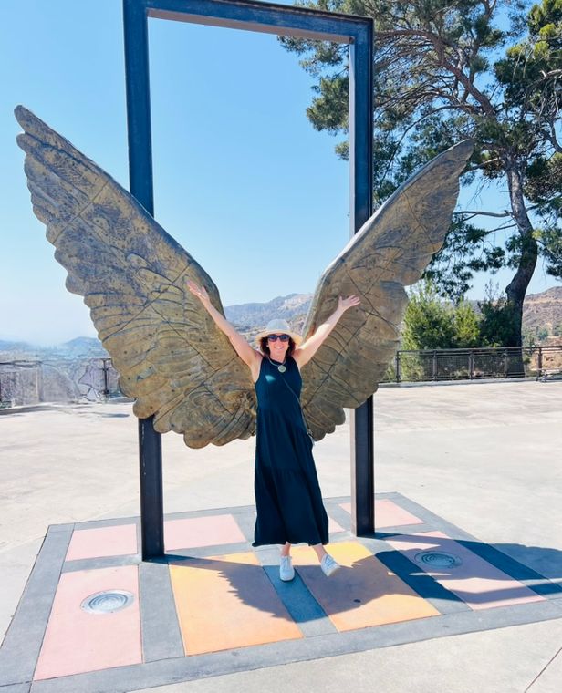 Claudia with her arms raised in front of a pair of sculpted bronze wings, the Hollywood sign in the distance