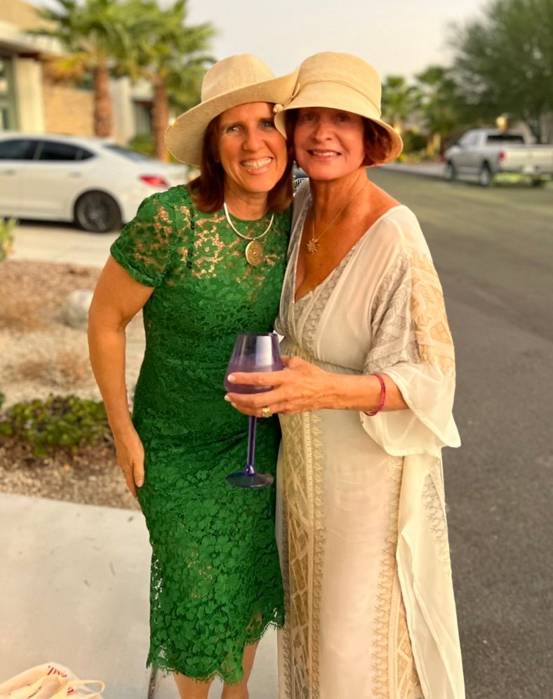 Claudia in a green lace dress and straw hat, arm in arm with a friend in a cream dress and hat, palm trees behind them
