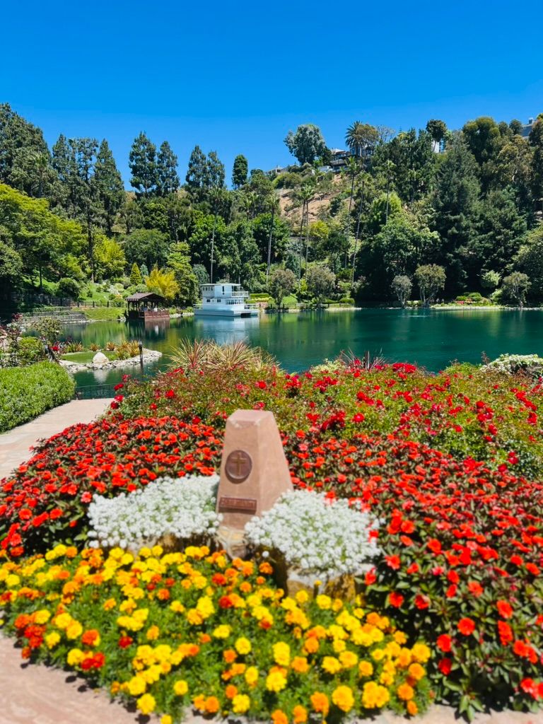 Red, white and yellow flower beds in front of the lake at Lake Shrine, a white houseboat and wooded hills beyond