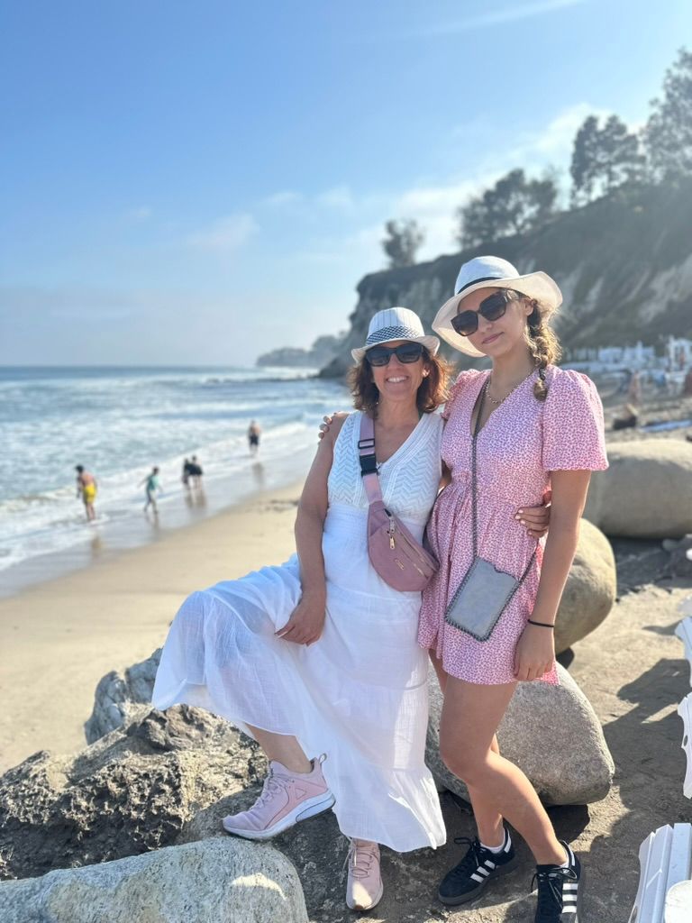 Claudia and her niece in sun hats, arm in arm on the rocks of a beach, waves and a cliff behind them