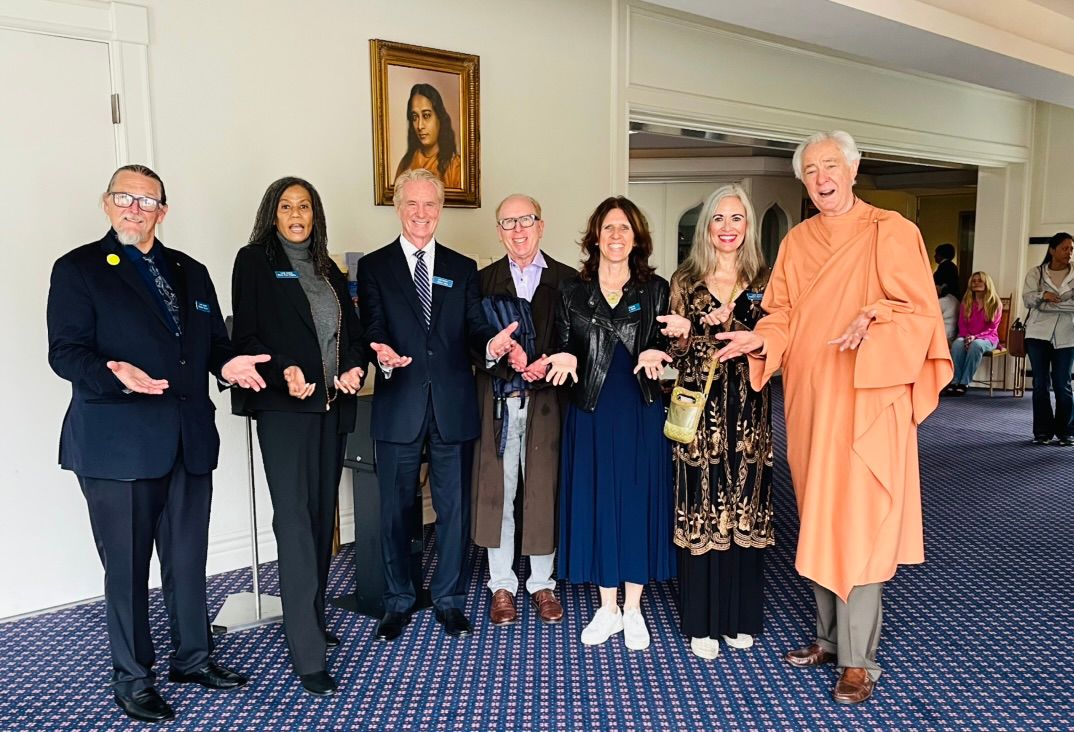 Claudia with a group, including a monk in an orange robe, all holding their palms up, a framed portrait on the wall behind them
