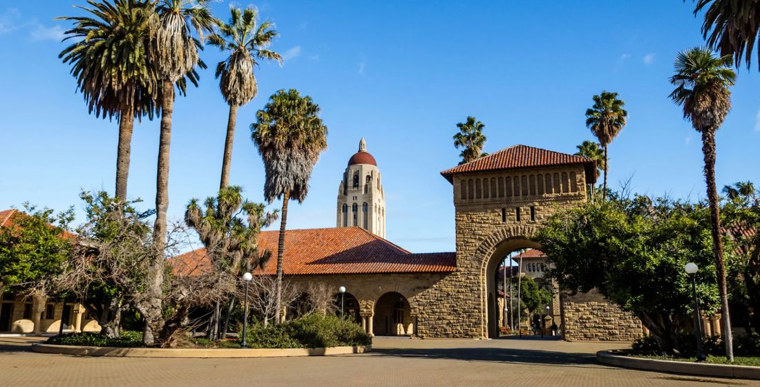 A sandstone archway on Stanford’s Main Quad, with Hoover Tower and palm trees against a blue sky