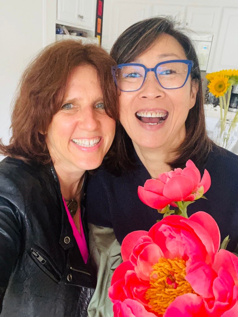 Claudia laughing in a selfie with a friend in blue glasses, big pink peonies in front of them