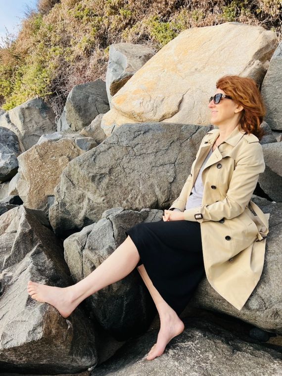 Claudia sitting barefoot on boulders in a trench coat and sunglasses