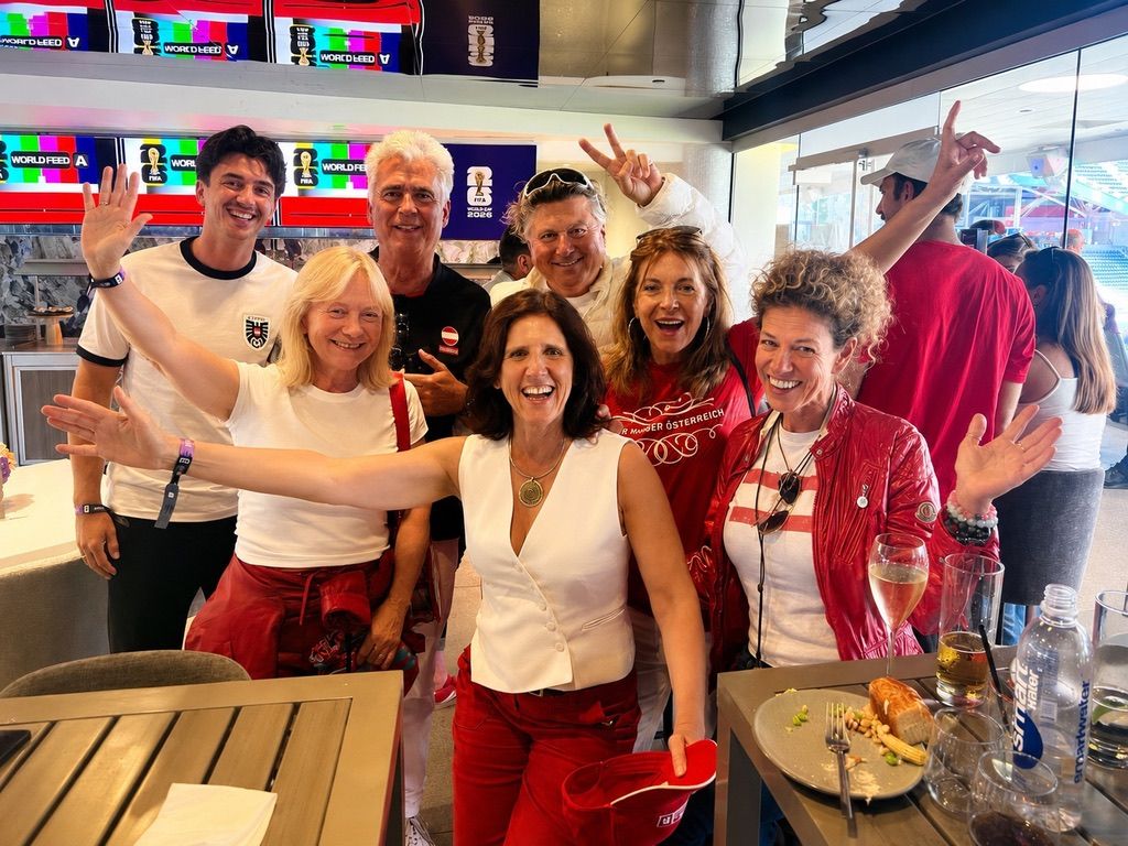 Claudia with her arms spread wide among a group of laughing Austria fans in red and white, in a stadium lounge at the 2026 World Cup